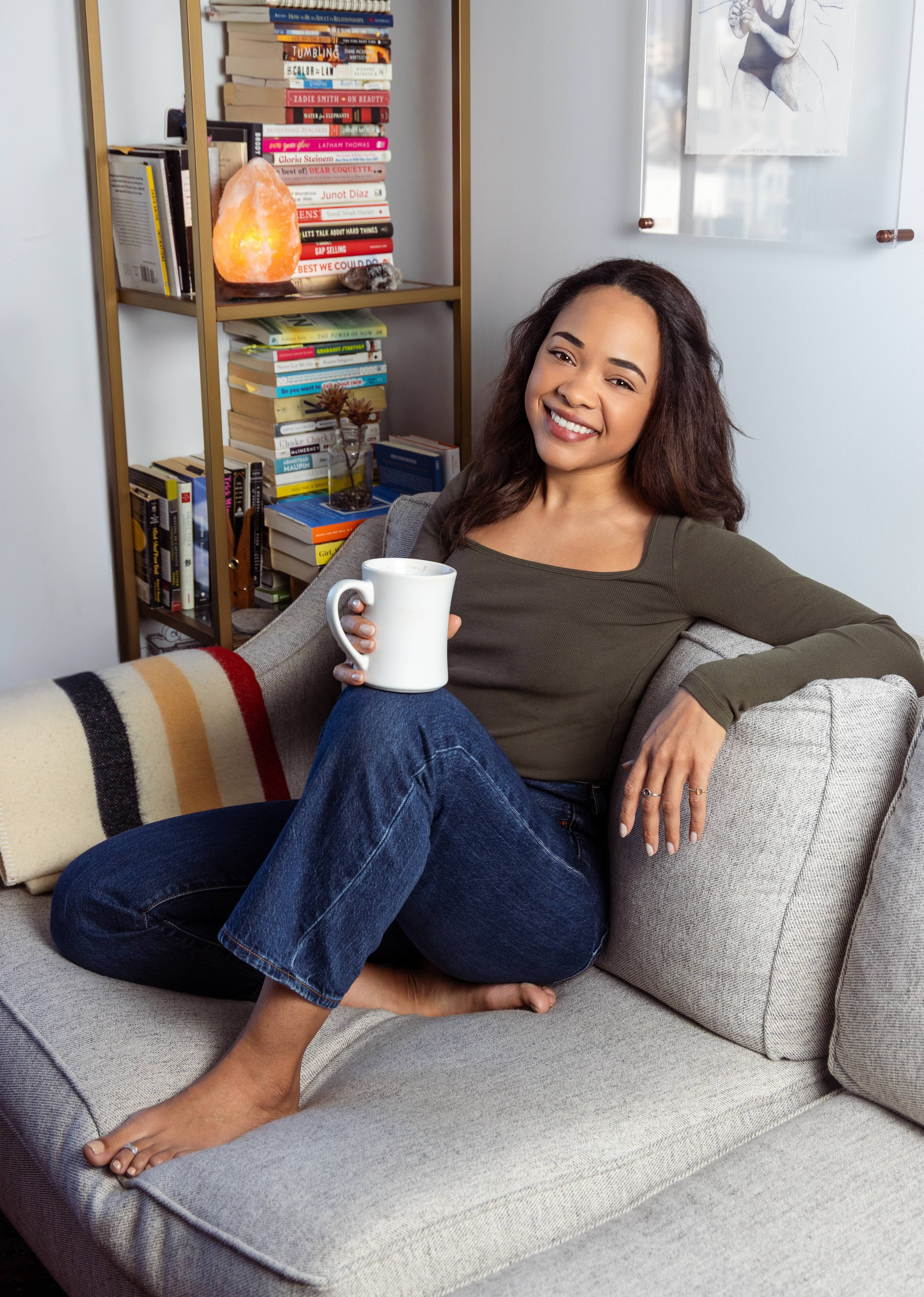Hélia Clamoungou seated on a couch with a mug, smiling, bookshelf behind her