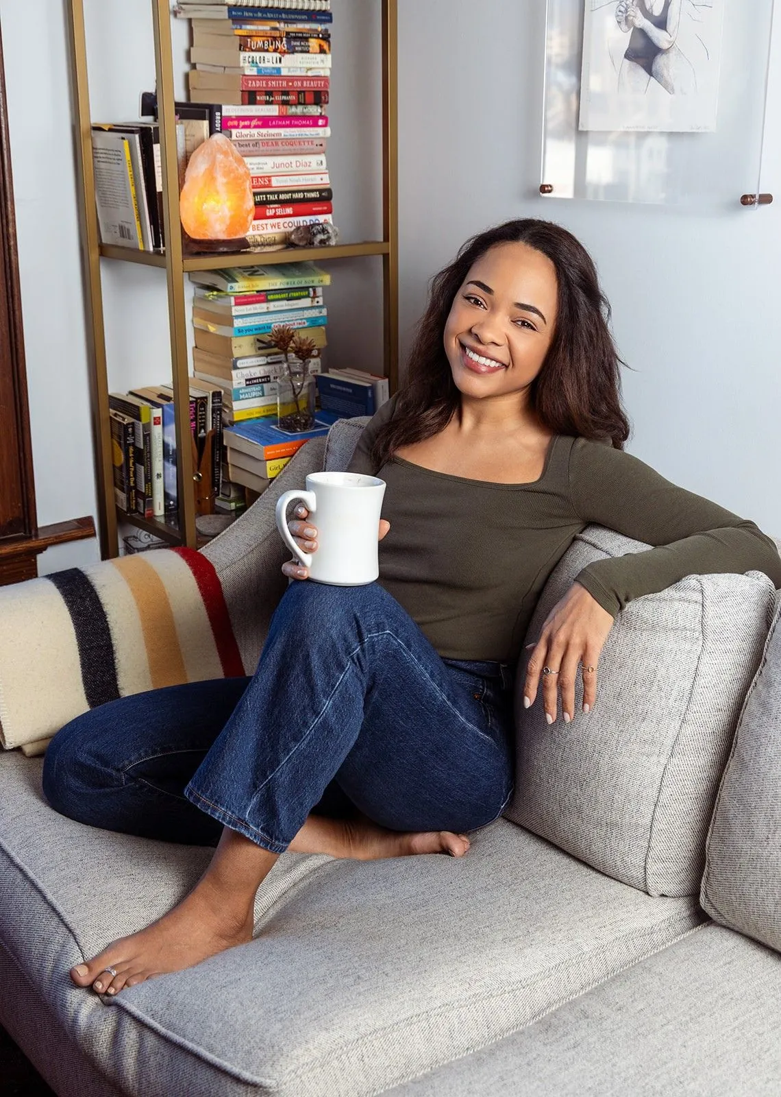 Hélia Clamoungou seated on a couch with a mug, smiling, bookshelf behind her